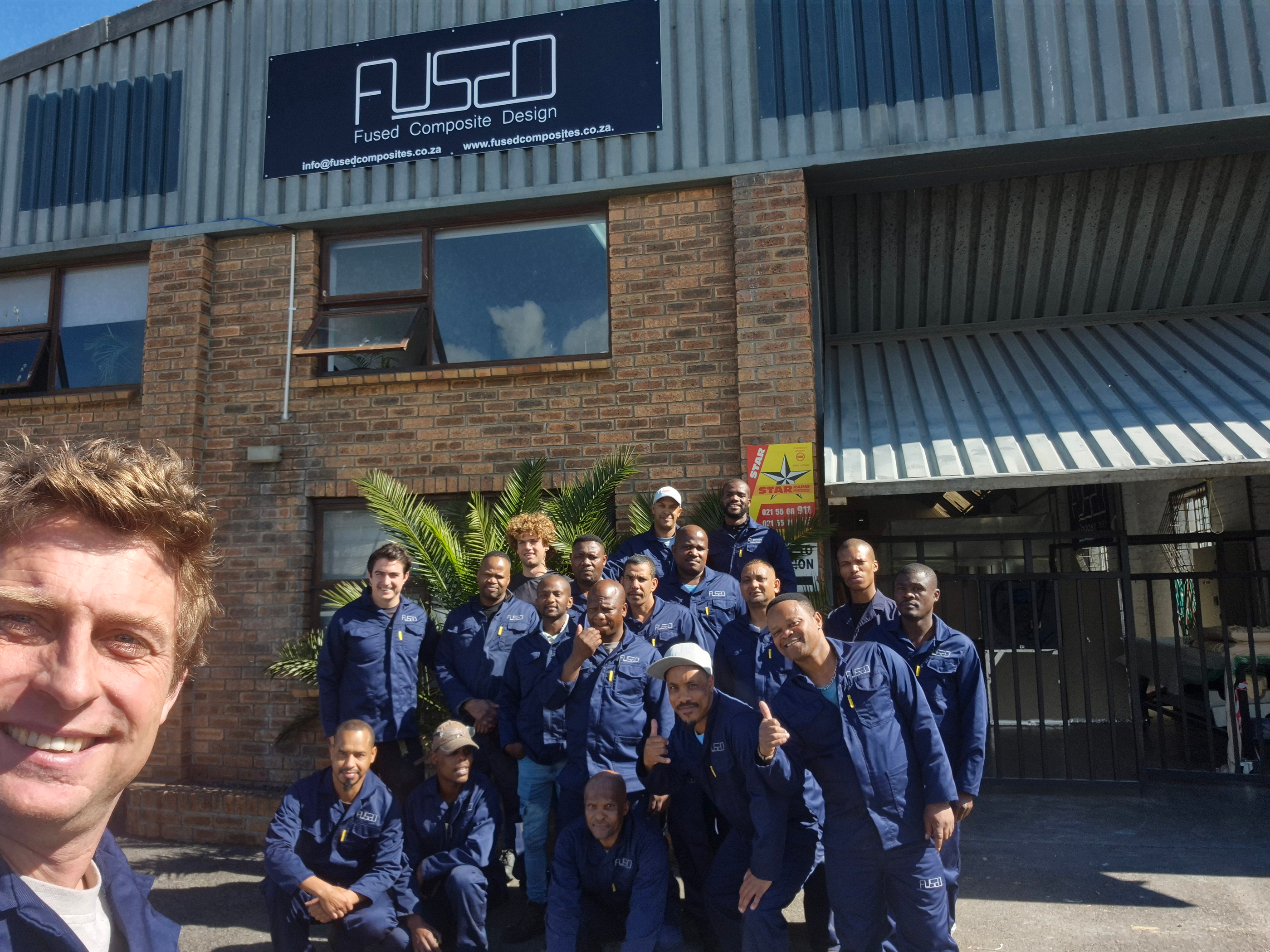 A large group of workers in navy overalls in front of a factory building with a Fused Composite Design sign, photographed by a smiling man in the foreground