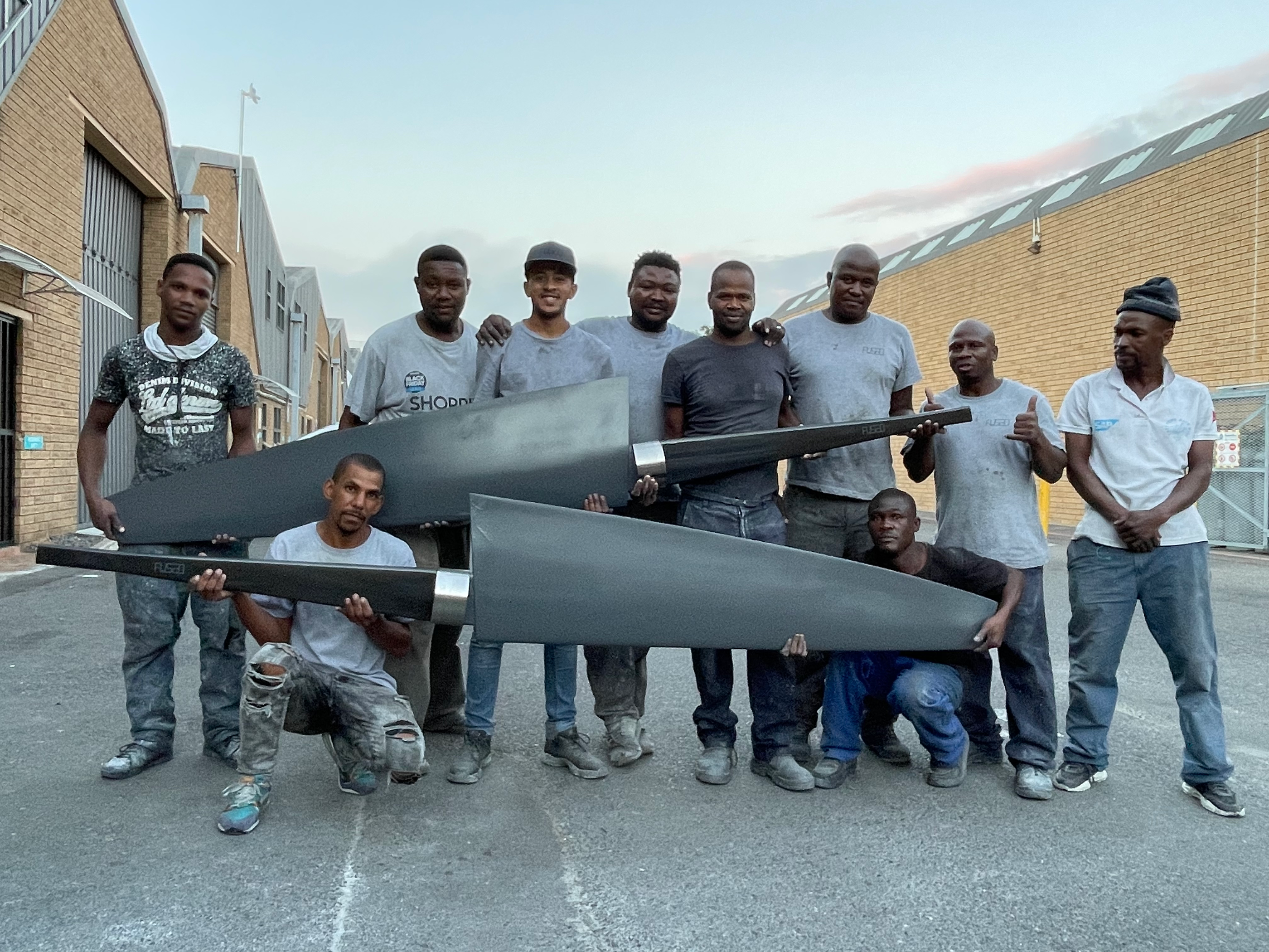 A group of ten workers outside a factory at dusk holding two large black carbon fibre foils