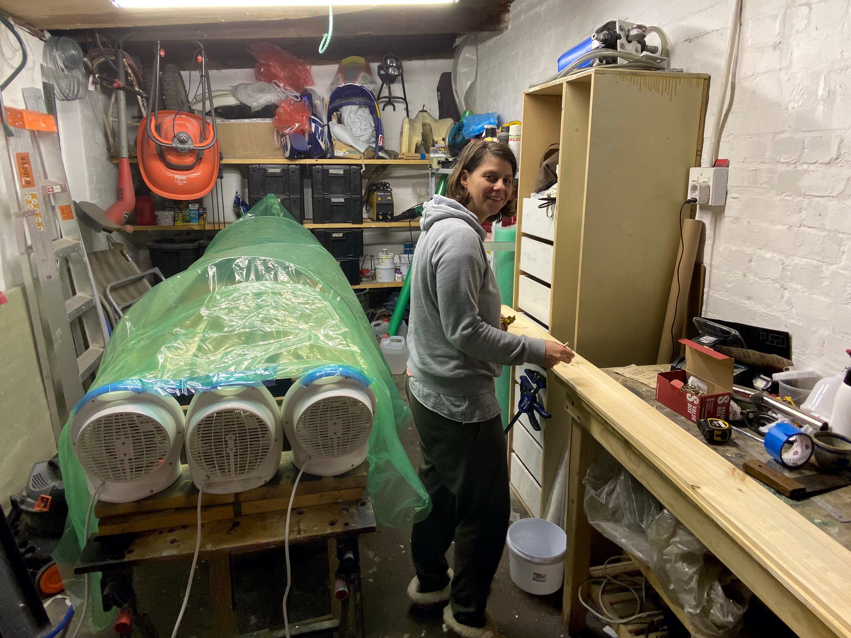 A long composite part under a green vacuum bag with three fan heaters beside it in a small garage workshop, a person standing at the bench