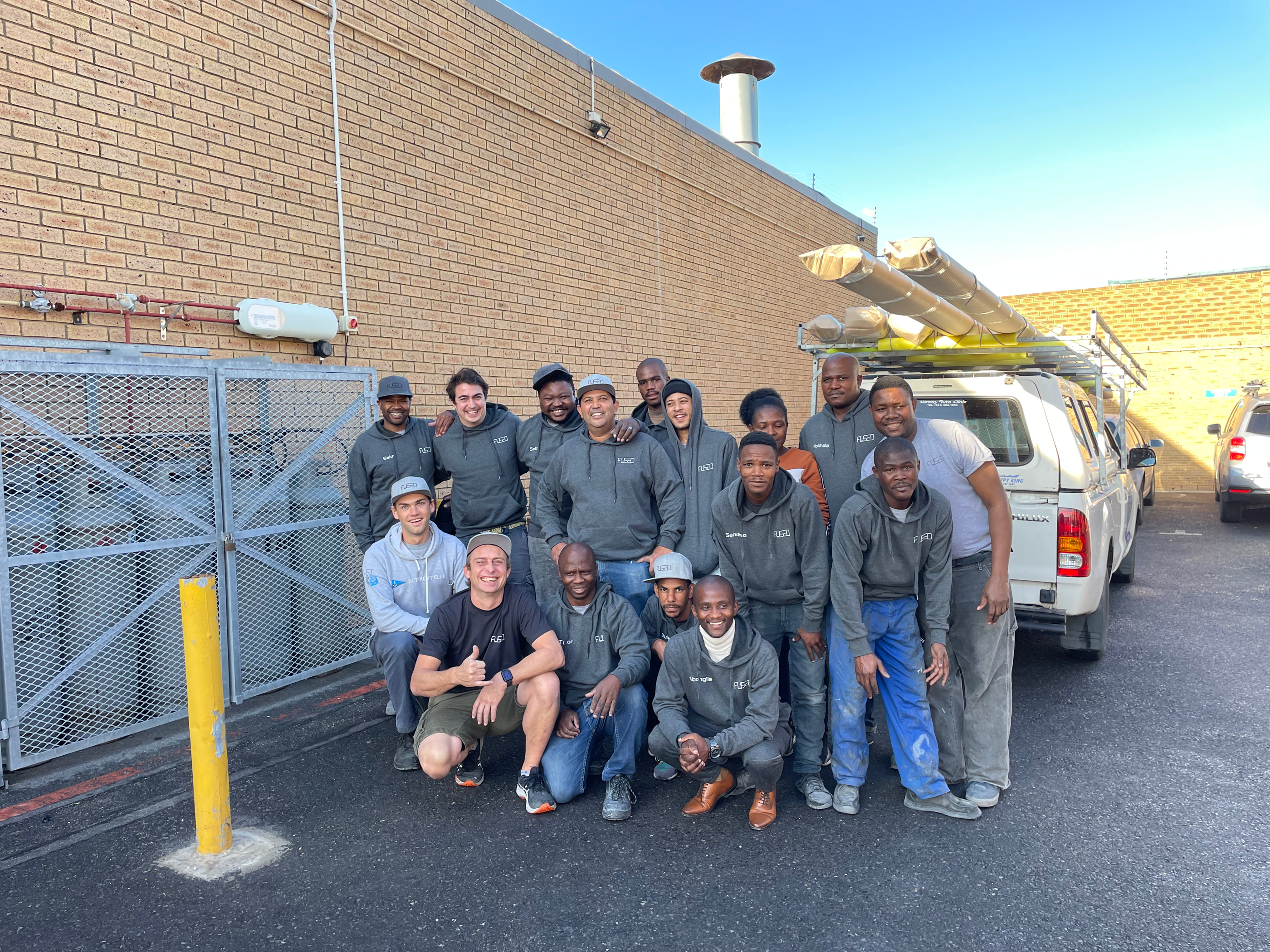 A large group of workers in matching grey hoodies posing in front of a factory gate and a loaded pickup