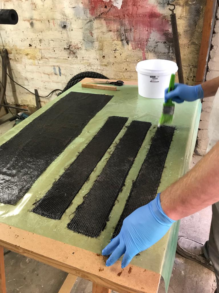 Gloved hands brushing resin onto strips of carbon fibre on a workbench