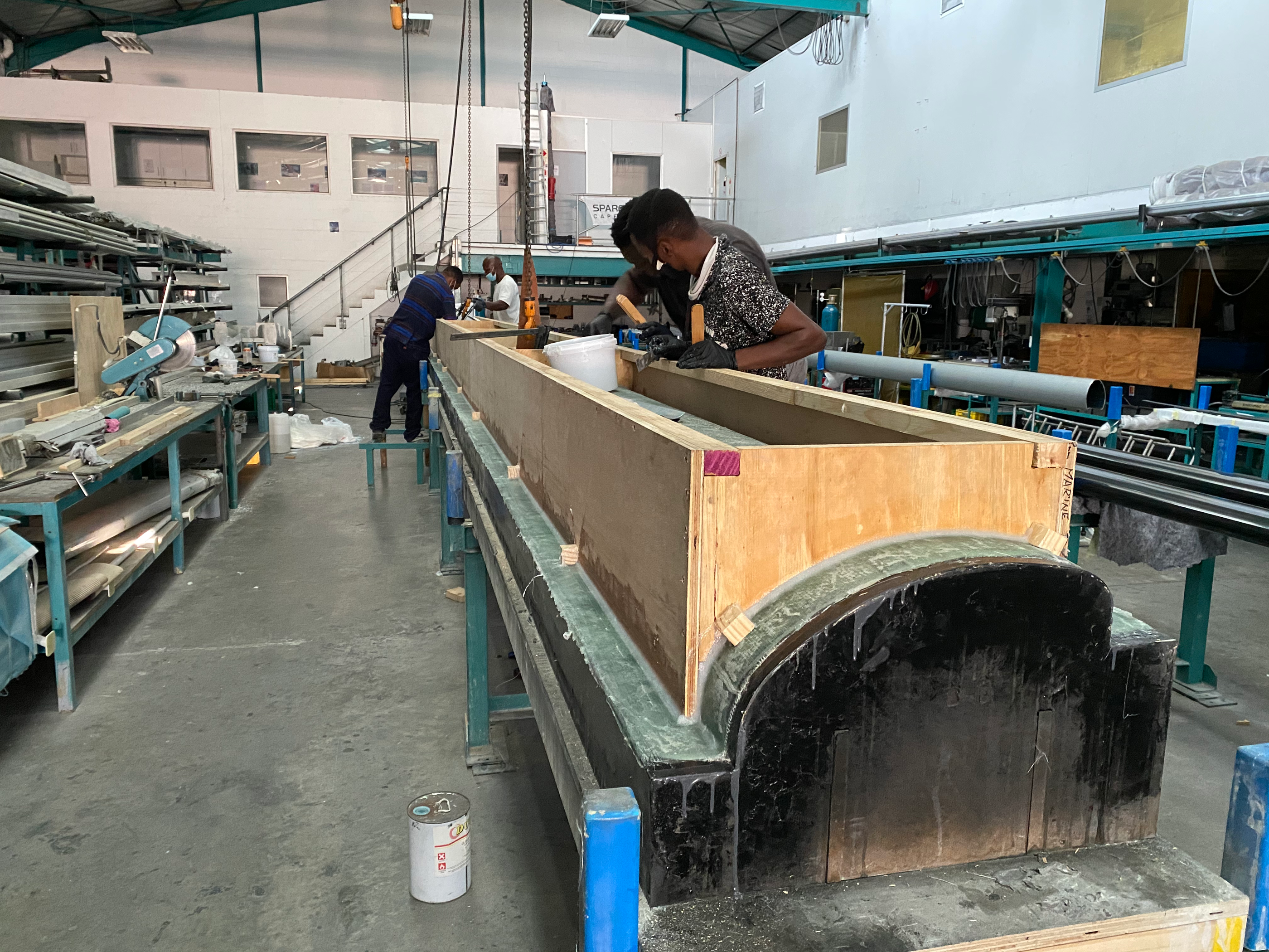 A long plywood spar mould on trestles in a busy composites workshop, technicians working in the background