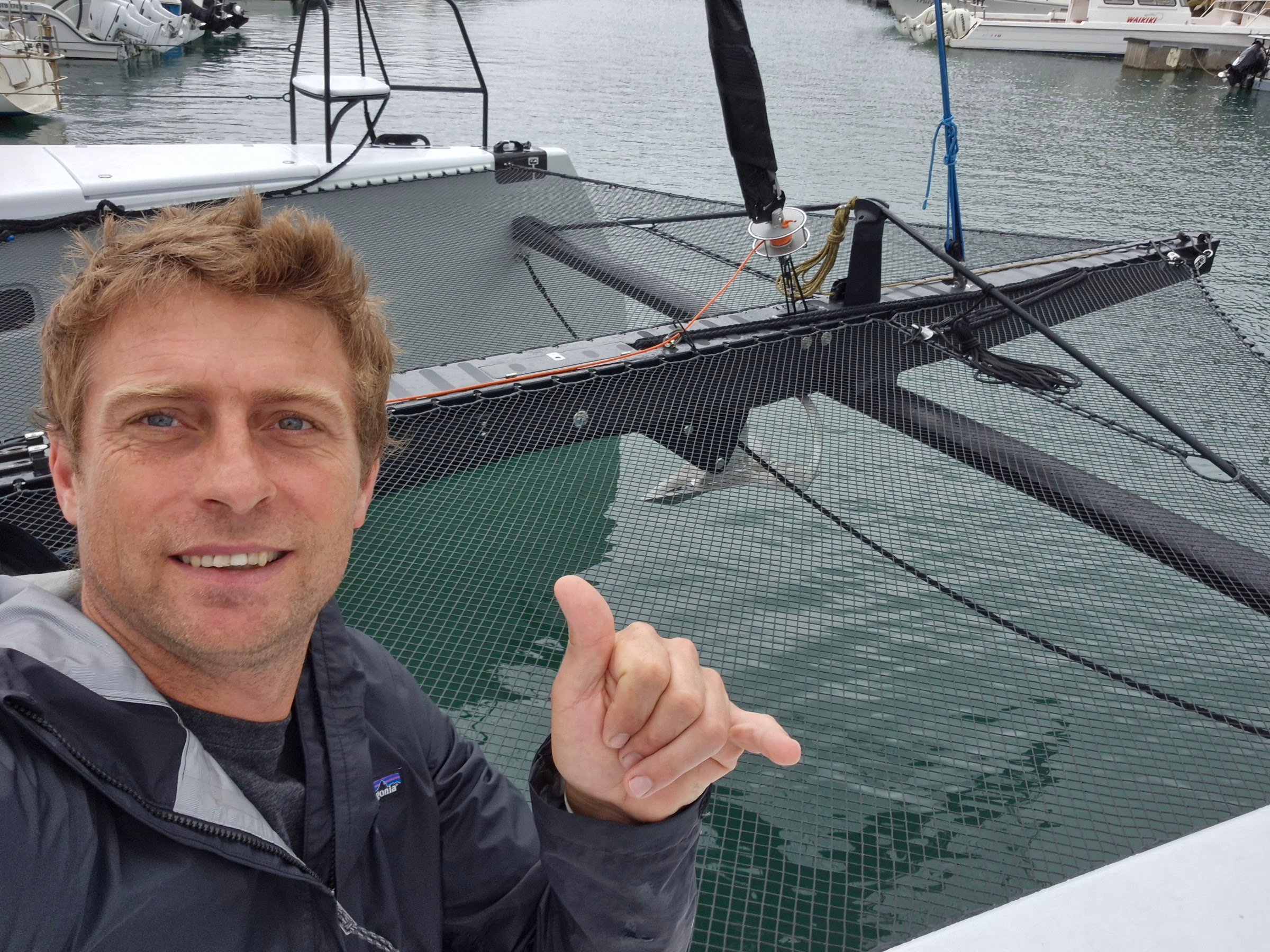 Catamaran afloat in a marina with forward beam, longeron and trampoline fitted, a smiling man in the foreground