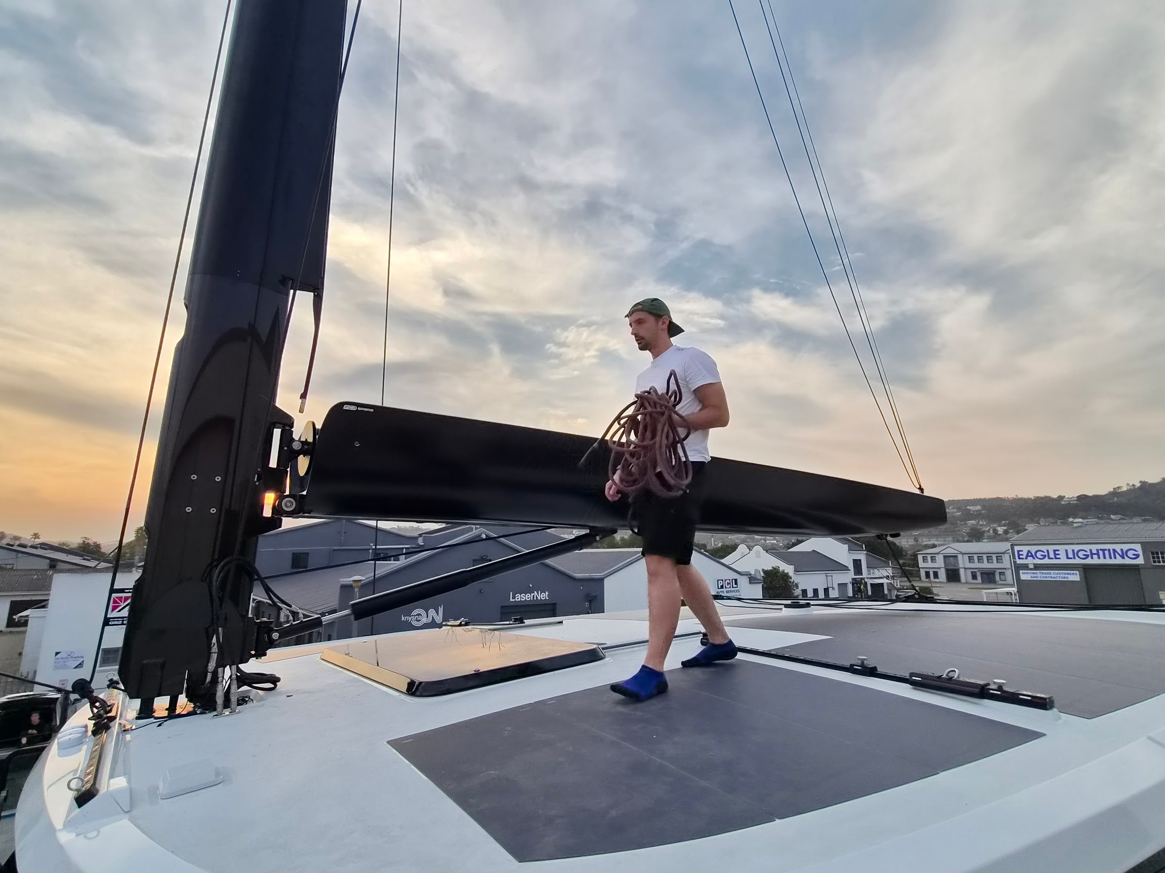 Technician carrying rigging line on a catamaran at dusk, fitted furling boom and mast behind