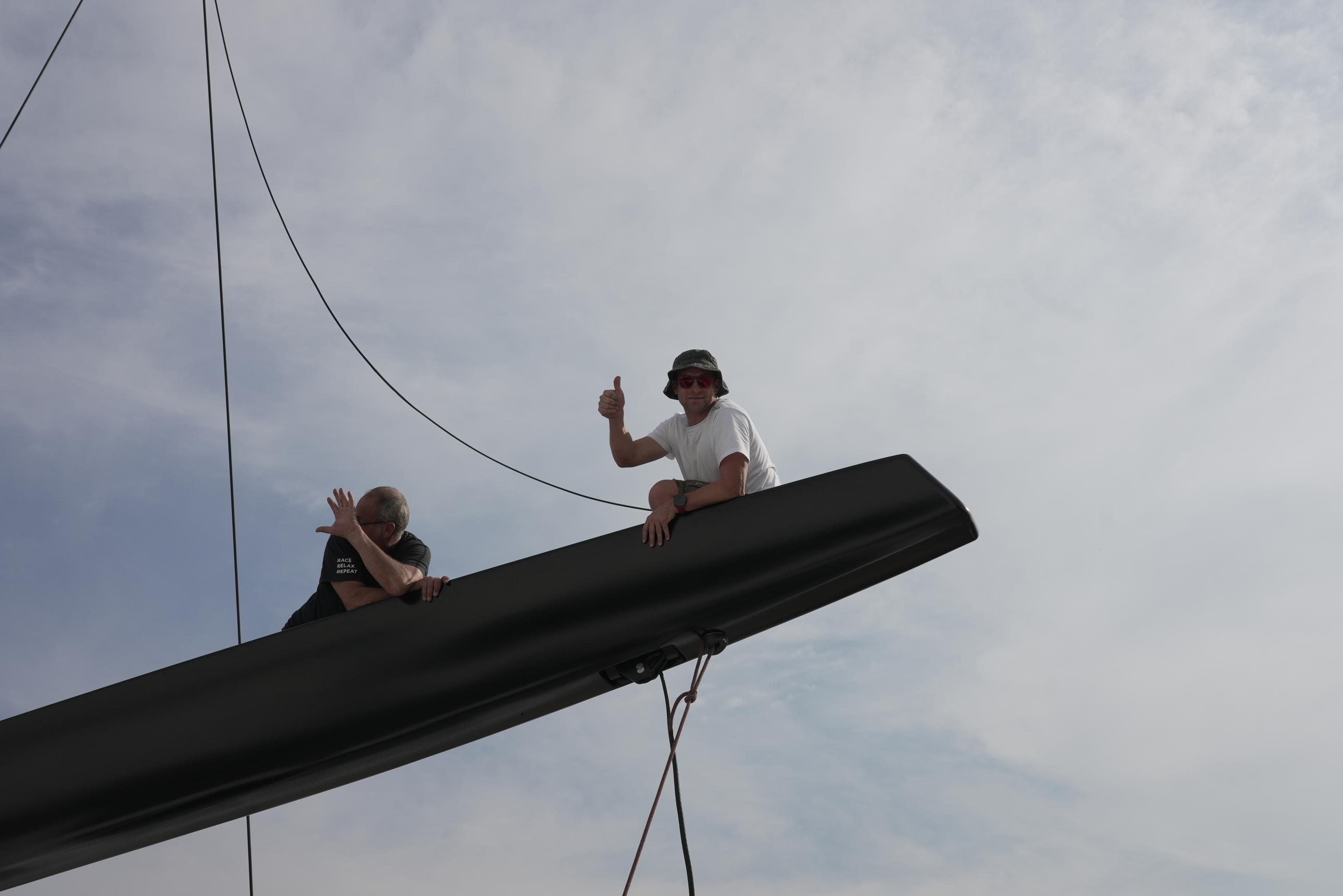 Two people sitting on the end of an installed carbon furling boom against the sky, one giving a thumbs up