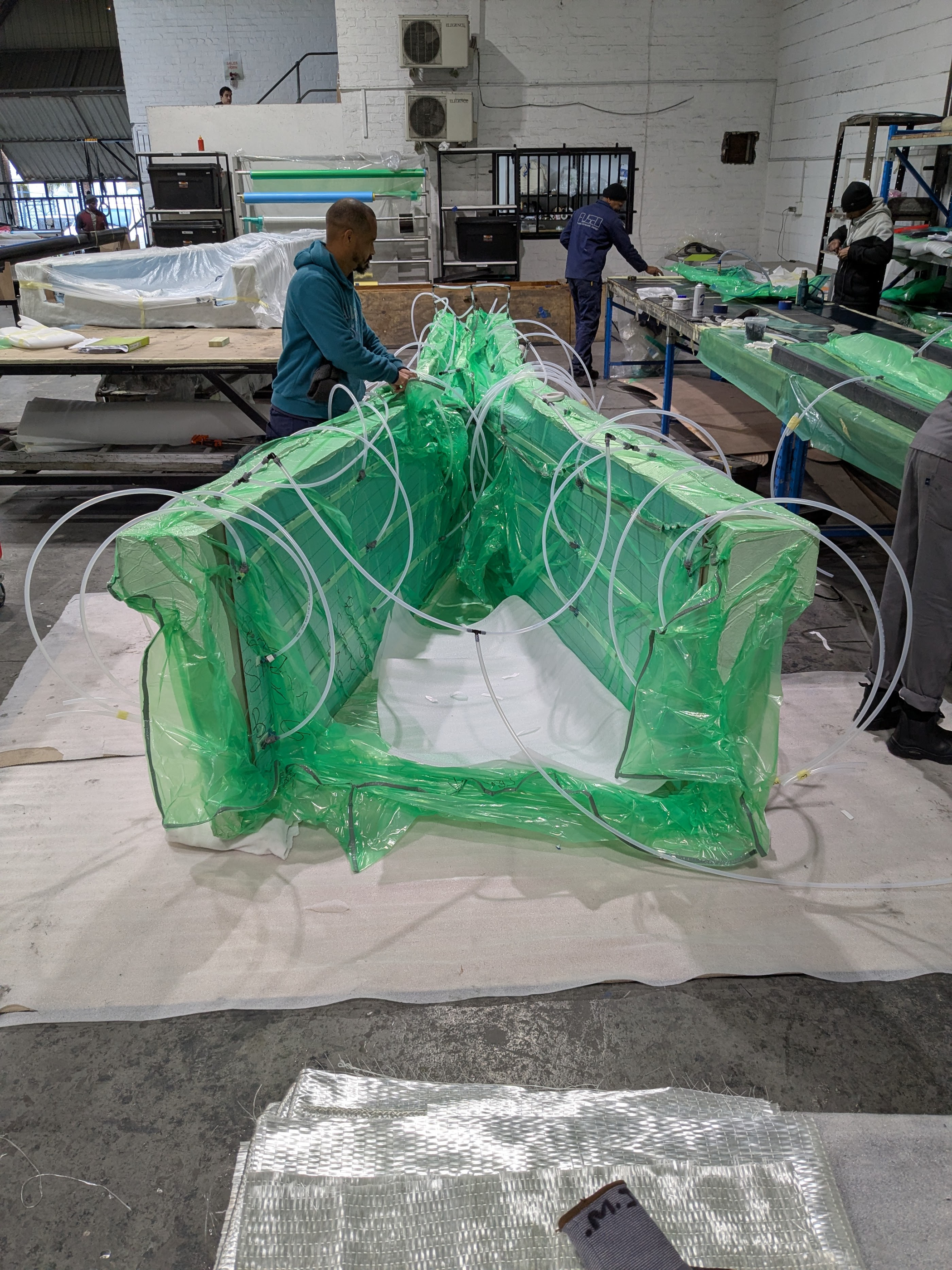 Composite part sealed under a green vacuum bag, technicians working around it in the factory.